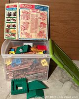 Plastic bin filled with Lincoln Logs wooden pieces and colorful plastic parts, original instruction sheet displayed behind.