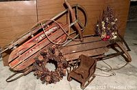 Photo showing two vintage wooden sleds, one with red paint, one unpainted, a pinecone wreath, dried floral spray, and small rusty sleigh grouped together on the floor.