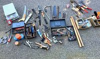 Wide overhead image showing multiple small hand tools, saws, pruning shears, paint brushes, tool boxes with assorted blades and small parts scattered on pavement.
