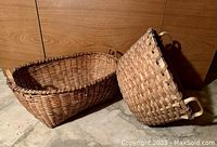 Photo showing two antique wood splint baskets, one rectangular and one round, both with handles, placed on the floor against a wood panel wall.