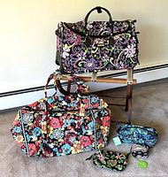 Two Vera Bradley patterned quilted bags, smaller makeup/toiletry bags, and a pill case arranged on floor and luggage rack