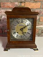 Front view of the walnut Warmink mantel clock with brass handle and feet, showing the clock face with Roman numerals and filigree accents.