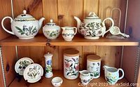 Wide view of shelf displaying assortment of ceramic items including teapots, canisters, plates, and tea strainer featuring botanical floral designs.