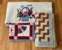 Three folded quilts on hardwood floor, top is appliqué floral, bottom left sampler, bottom right Rail Fence quilt.