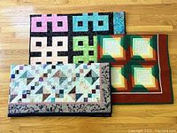 Three medium-sized quilts displayed on wooden floor: one folded quilt with pastel and floral patterns; a smaller quilt with warm earthy hues; and a black background quilt with bright multicolor key-like patterns and blue-green border.