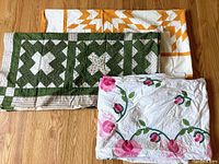 Three folded quilt tops: green-white Crossroads variation, pink rose appliqué vine, and gold star top behind.