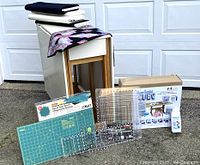 Front view of entire lot arranged outdoors against garage door; includes cutting table, quilts, storage bin, cutting mat, rulers and fabric bolts
