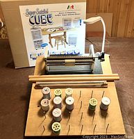 Full setup showing Durand pleating machine on wooden base with spools of thread, two wooden dowels in front, Cube Super Satchel organizer box behind, and white task lamp attached.