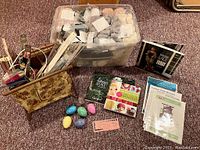 Wide shot showing wooden folding yarn caddy, plastic bin of yarn skeins, knitting pattern and instructional books, and stone darning eggs on carpeted floor