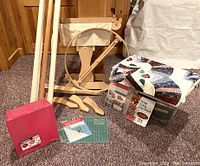Wide shot showing the wooden quilting hoop on floor stand, wooden rods, fabric bin, cutting mats, templates, quilting book and additional tools. The quilting hoop is about 30 inches in diameter with a natural wood finish floor stand and a canvas holder attached.