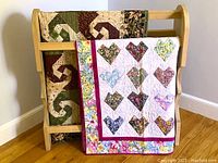 Two quilts displayed on a wooden quilt stand, one colorful 'Crazy Hearts' with heart shapes, one patterned 'Wonky Monkey Wrench' quilt in earth tones.