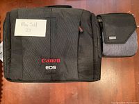 Front view showing black Canon EOS camera case and smaller gray/black Think Tank camera case side by side on wooden surface.