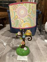 Front view of the lamp showing the tree trunk with two owls, flowers, green base, and patterned lampshade with pink trim.