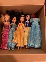 First photo showing five Disney princess dolls dressed in various signature gowns placed side by side in cardboard box.