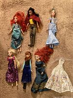 Seven Disney princess dolls laid out on beige carpet with different colored dresses and hairstyles plus one separate new princess wedding dress.