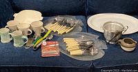 Varied kitchenware items displayed including ceramic mugs and cups, plastic platters, metal colander, small pottery pieces, and cutlery with ivory handles.