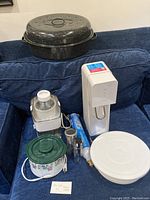 Graniteware turkey roaster, white juicer, ceramic crockpot, Sodastream, gas cartridge, salt shaker, pepper grinder, and cake decorating stand shown on a blue couch.