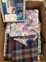 Box containing a Conair electric clothing shaver in box and folded fabric in purple floral and plaid patterns.
