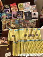 Top-down view of all books laid out flat showing mostly vintage Nancy Drew yellow spine paperbacks and assorted mystery and fiction books on a carpeted floor