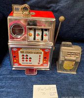 Two vintage one armed bandit coin banks shown side by side. Larger bank has red top and metallic body with classic slot machine design. Smaller bank is silver with 10¢ marking and jackpot display.