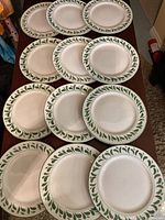 Top view of all 12 Williams Sonoma plates arranged in two rows on a brown surface, showing the green leaf border design and slight variation in lighting reflecting minor wear.