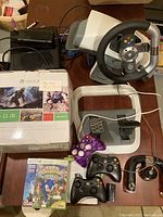 Overall lot showing Xbox 360 console, controllers (standard and Rock Candy), racing wheel with foot pedals, original box, and some game discs.