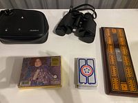 Photo showing Celebrity brand binoculars with black carrying case, Gustav Klimt-themed playing cards, and wooden dominoes board arranged on a white surface.