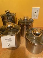 Photo of four stainless steel canisters with glass lids and stainless steel handles arranged on a countertop in front of a yellow-orange wall and electrical outlet.
