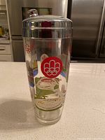 Front view of vintage Montreal 1976 Olympics cocktail shaker showing Olympic rings, Montreal 1976 logo, and colorful city and Olympic venue graphics.
