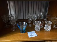 Overview of 18 crystal and glass ware items on wooden shelf including cut crystal wine glasses, Holmegaard Princess glasses, two Longchamp vases, and a small blue glass bowl.