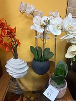 Three faux orchid flower arrangements on a table against yellow wall: white orchids in black and beige pots and orange orchids in a white spiral vase.