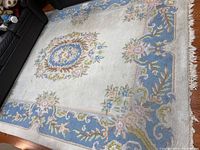 Top view of handmade wool area rug with floral design and blue border, showing fringe along the edge, placed on floor next to black furniture.