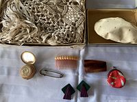 Photo showing the hand-woven shawl with macramé fringe, vintage compact, hair combs, safety pin, earrings, and small red coin purse on white surface.