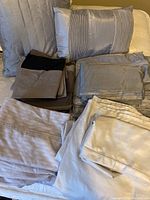Overall view of four sets of double bed sheets in neutral tones with two accent cushions in silver satin and quilted fabric.