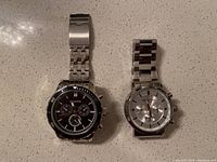 Two men's wristwatches placed side by side showing silver bands and round faces, one with a white dial, the other black.