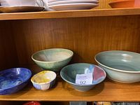 Six assorted pottery bowls displayed on wooden shelf showing variety in size, shape, and glaze color.