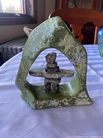 Full view of soapstone sculpture showing arch frame and centered Inukshuk figure on white cloth with background window and furniture.