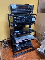 Black metal stereo stand holding several pieces of audio equipment such as a tuner, amplifier, DVD player, and VCR, photographed against a yellow wall with a wooden floor.