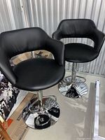 Photo showing two black padded adjustable bar stools with curved backrests and chrome bases. Stools have footrests and hydraulic height adjustment.