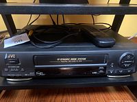 Front view of black JVC HR-DD74OU VHS player showing the power button, AV inputs, menu, and channel controls with remote and cables on top.