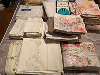 Folded assorted bed linens including embroidered white pillowcases, plain sheets, and floral patterned sheets.