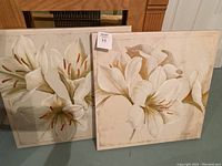 Two complementary canvas art pieces featuring white lilies with brown and beige tones on a cream background, both roughly 24 inches square.