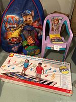 Photo showing the Paw Patrol tent, sports backpack with balls, two small plastic camping chairs, and the boxed gigantic step piano.