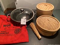 Hand hammered wok, glass lid, bamboo steamer baskets, whisk, and red cloth bag all shown together on floor.