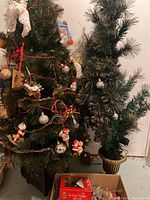 Two artificial Christmas trees side by side, one decorated with assorted ornaments and red beads, the other undecorated in a gold-colored pot stand.
