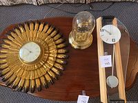 Overview of the three clocks on wooden table including Syroco sunburst, quartz dome clock, and wooden pendulum clock