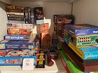 Photo showing a collection of games and puzzles including board games and boxed puzzles stacked on shelves, some titles visible such as Yahtzee, Jenga, Payday, and puzzle brands like Springbok and WS.
