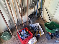 Photo showing multiple rakes, shovels, crates filled with outdoor chemical bottles and hand garden tools, a green watering can on floor