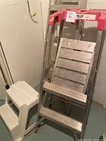 View of white plastic step stool next to aluminum 4-foot platform ladder with red top and an aluminum stepladder behind.