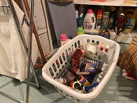 Photo showing laundry basket filled with assorted items including cleaning supplies, detergent bottles in background, part of ironing boards visible